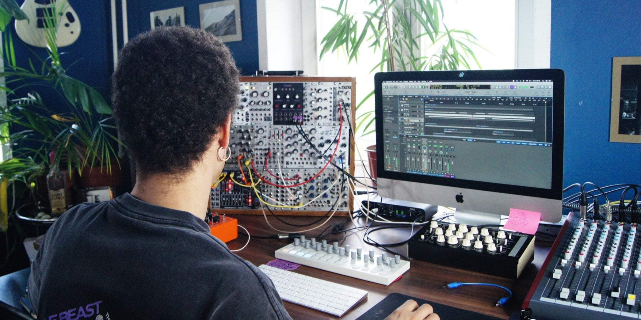 Person working at a computer with audio equipment in a room with plants and a guitar on the wall.